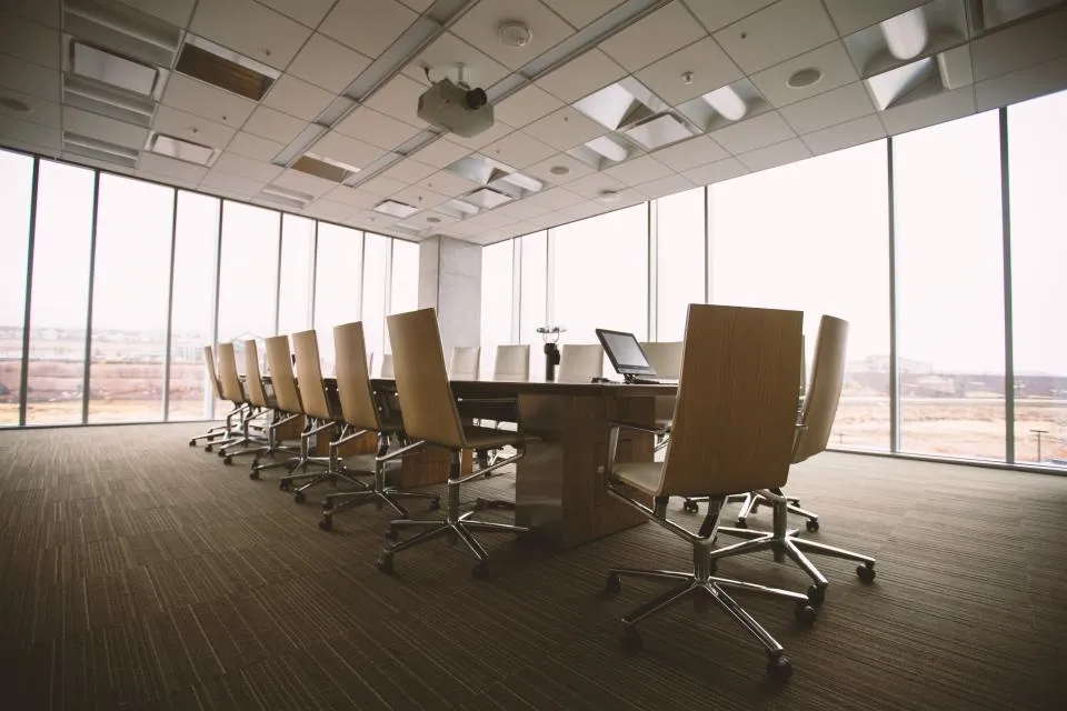An empty boardroom with a long table and rows of chairs facing tall windows