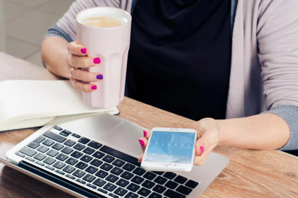 A person at a laptop holds a coffee cup and a smartphone
