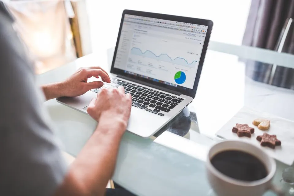 A person works on a laptop showing line charts and a pie chart, a cup of coffee beside it