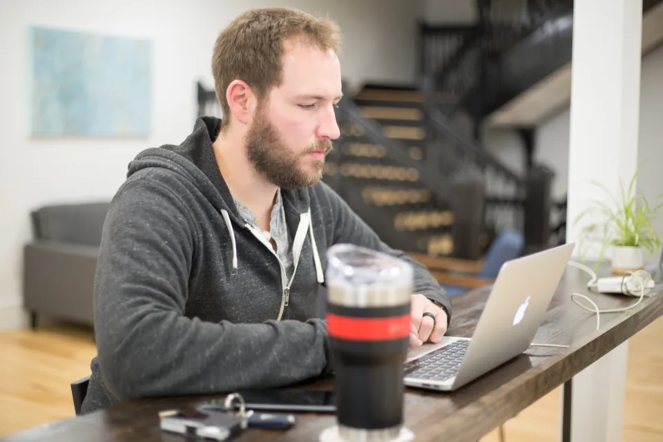 A man in a grey hoodie works on a laptop at a wooden desk