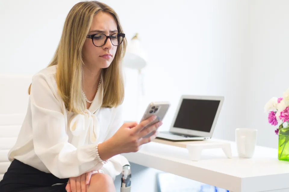 A woman in glasses at a white desk checks her phone, a laptop open beside her
